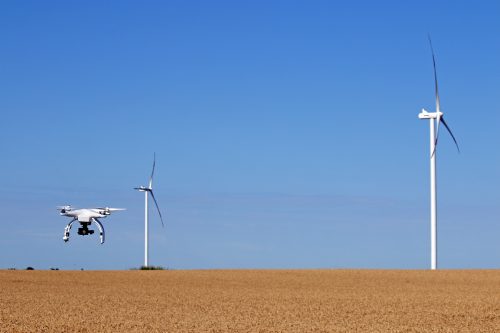 Quadcopter drone flying in a clear blue sky on a sunny day, with wind turbines in the background over a golden field, highlighting modern agriculture and renewable energy.
