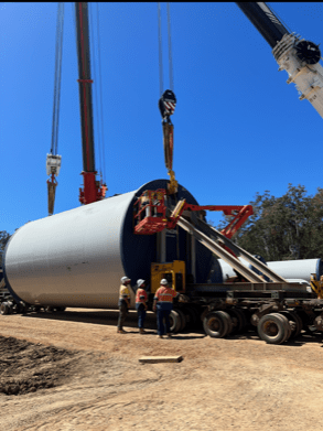 Construction crew at a renewable energy site hoisting a large wind turbine section with cranes against a clear blue sky, highlighting industrial teamwork.