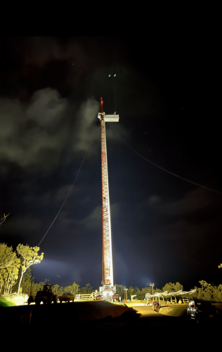 Nighttime view of a lit wind turbine construction site with a clear spotlight on the towering structure, showcasing the scale of wind energy projects against a dark, cloud-streaked sky.