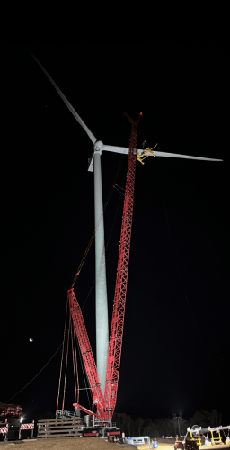 Nighttime installation of a wind turbine with a large red crane, illuminated by artificial lighting, against a dark sky, showcasing renewable energy development.