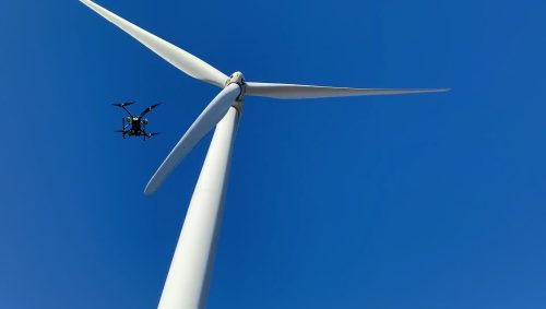 Drone conducting inspection near the hub of a wind turbine with spinning blades against a clear blue sky, emphasising high-tech maintenance in renewable energy.