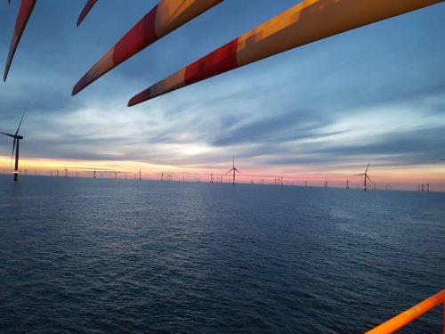 Offshore wind turbines captured at sunset with a dramatic sky, viewed from a perspective with turbine blades in the foreground, highlighting sustainable ocean energy.